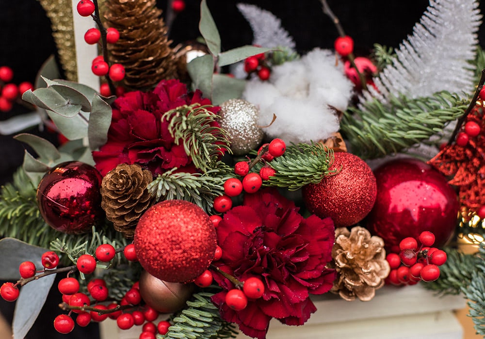 Close-up view of holly, pine cones, ornaments and red blooms in a holiday-themed arrangement